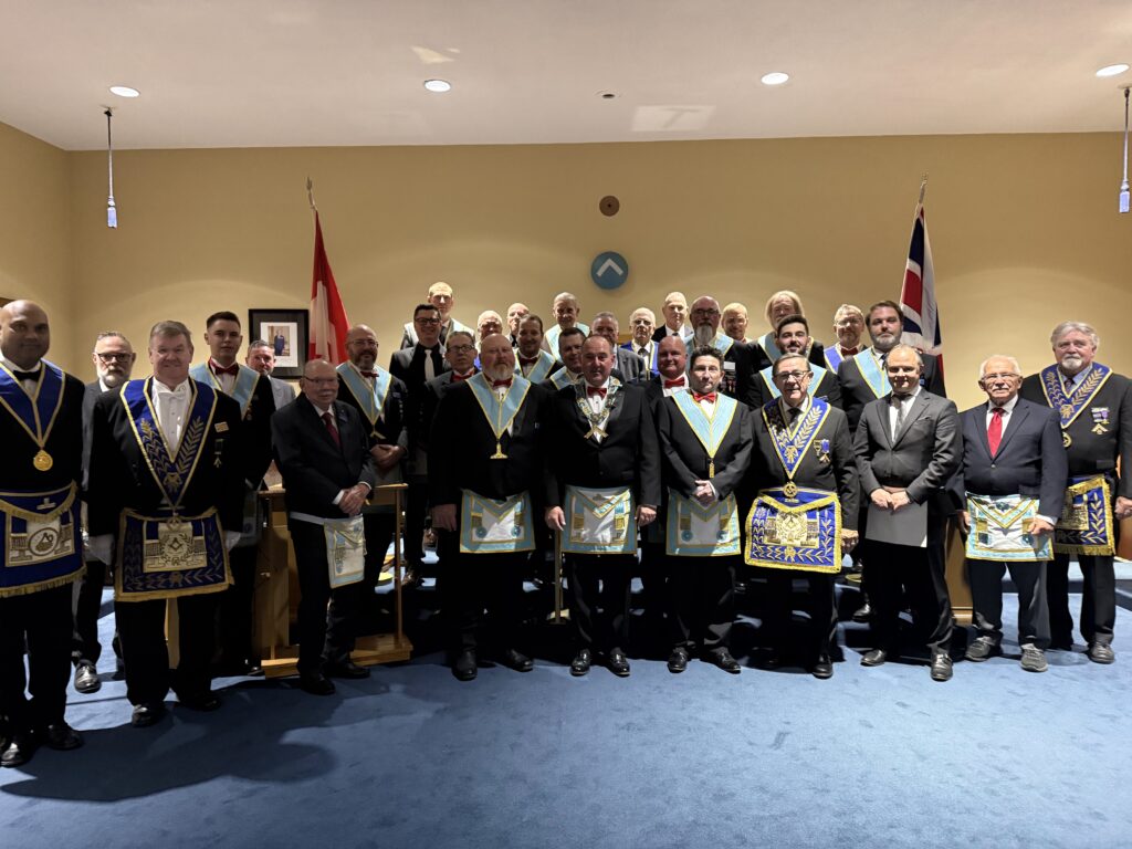 A group photo of lodge members standing together in a ceremonial room during an installation event. Several members wear Masonic regalia—including aprons, collars, and medals—while others are in formal suits. Two national flags are displayed behind the group, and the setting features a blue carpet and warm lighting.