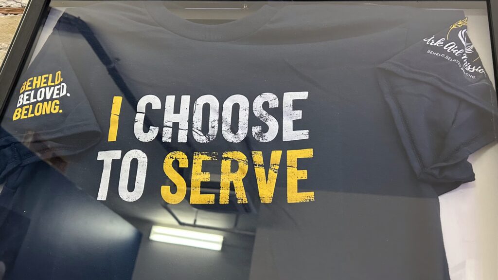 Framed t-shirt displayed in a shadow box at Ark Aid Mission, preserved as a meaningful piece representing community support, outreach, and the organization’s impact on those in need.