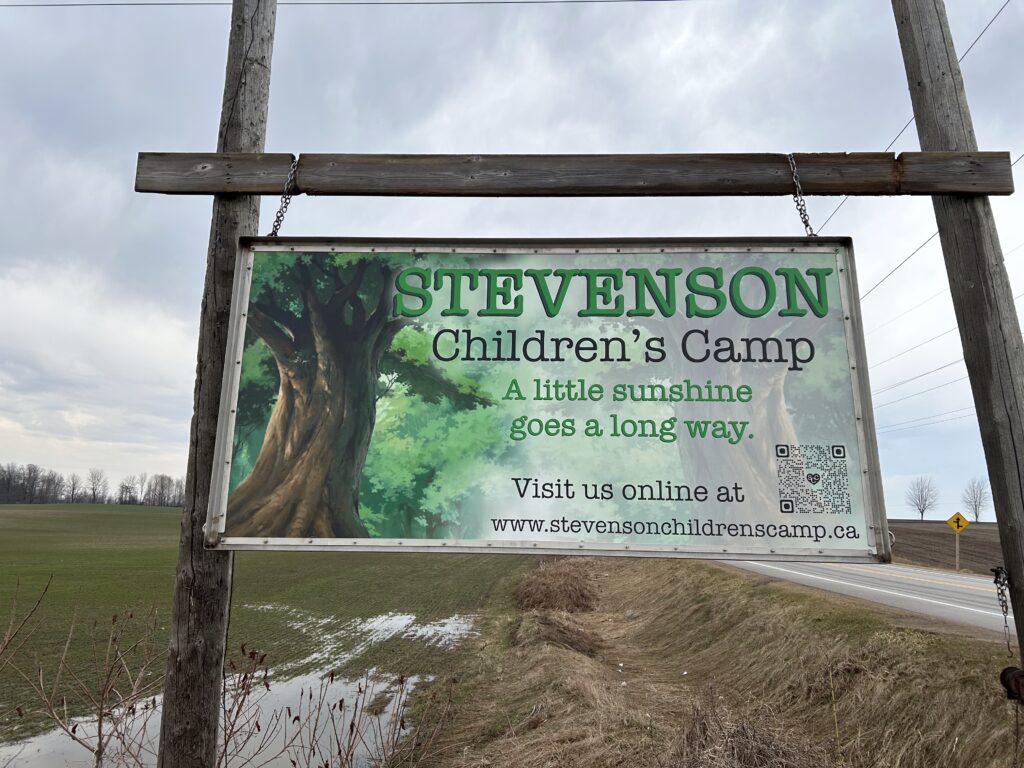 Wooden camp sign at the entrance of a rustic campsite surrounded by natural outdoor scenery, marking the camp location for visitors.