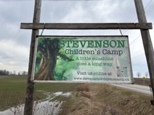 Wooden camp sign at the entrance of a rustic campsite surrounded by natural outdoor scenery, marking the camp location for visitors.