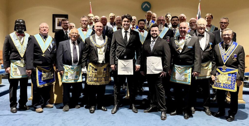 Group of Freemasons in formal attire and aprons gathered inside a lodge room after a ceremony.