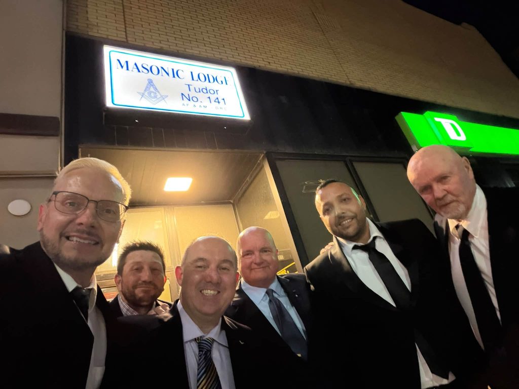 Freemasons from the Tuscan 195 posing outside Tudor Lodge No. 141 in Mitchell, Ontario, beneath the illuminated Masonic Lodge sign.